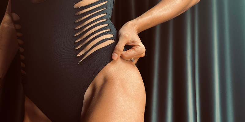 Person wearing a black swimsuit with a unique design, adjusting the strap. With a nice spray tan on.
